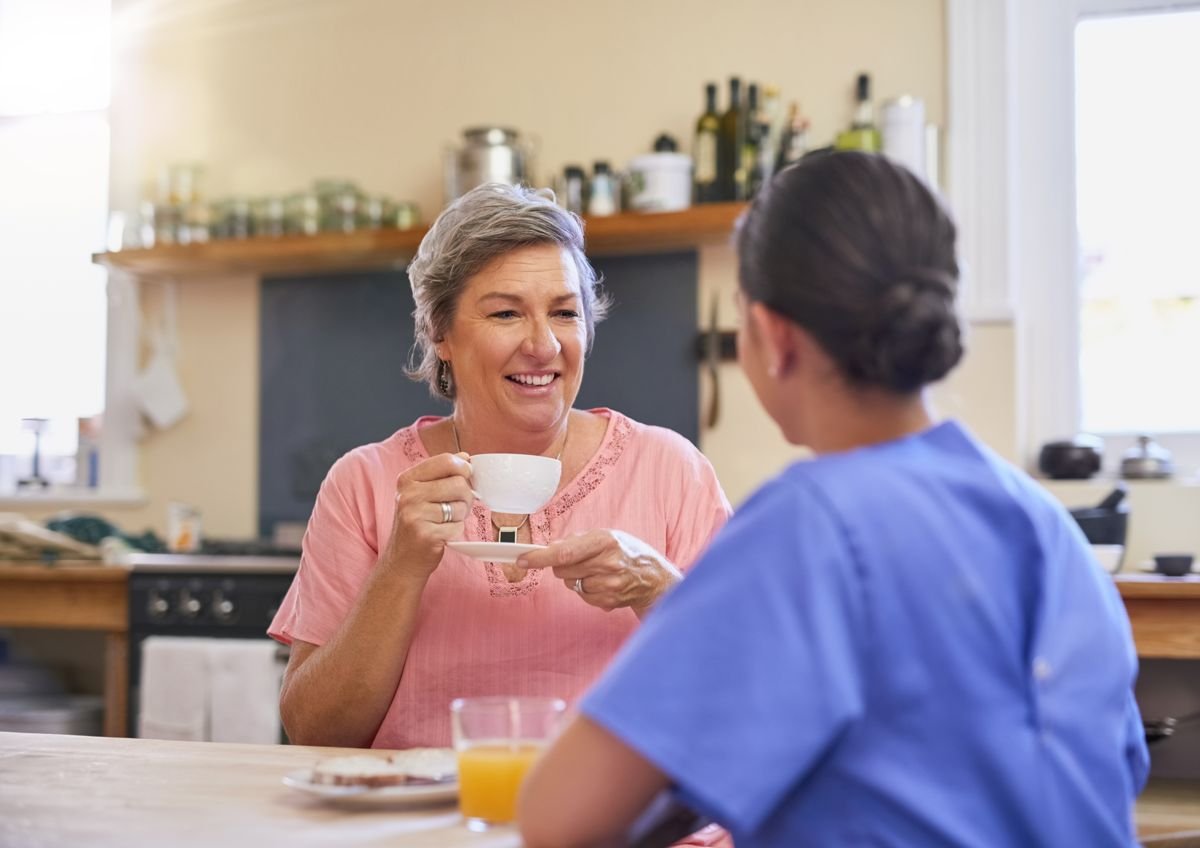 Una cuidadora y una mujer mayor conversan tomando un té en la cocina